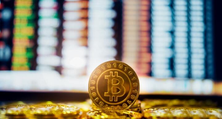 A gold coin stamped with a bitcoin logo in front of a computer screen